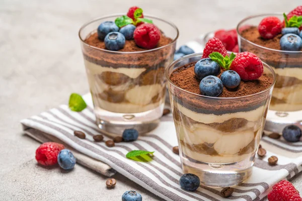 Sobremesa tiramisu clássico com mirtilos e framboesas em um copo na toalha de cozinha no fundo de concreto — Fotografia de Stock