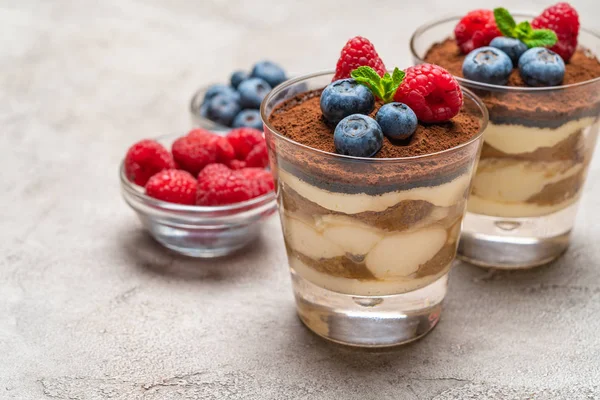 Klasszikus tiramisu desszert áfonyával és málna egy pohár és tálak bogyókkal a beton háttér — Stock Fotó