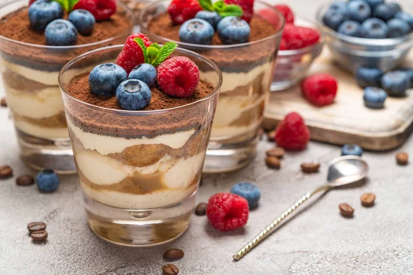 Klassisches Tiramisu-Dessert mit Blaubeeren und Himbeeren im Glas und Schalen mit Beeren auf Betongrund — Stockfoto