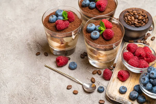 Klasyczny deser Tiramisu z jagodami i malinami w szklance i miski z jagodami na betonowej tle — Zdjęcie stockowe