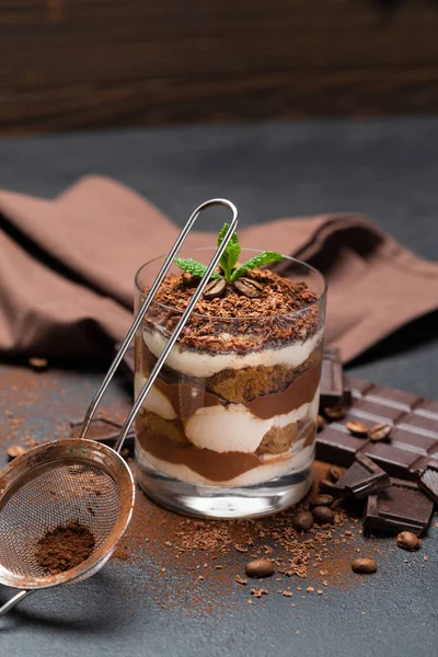 Sobremesa tiramisu clássico em uma xícara de vidro, filtro e pedaços de chocolate no fundo de concreto escuro — Fotografia de Stock