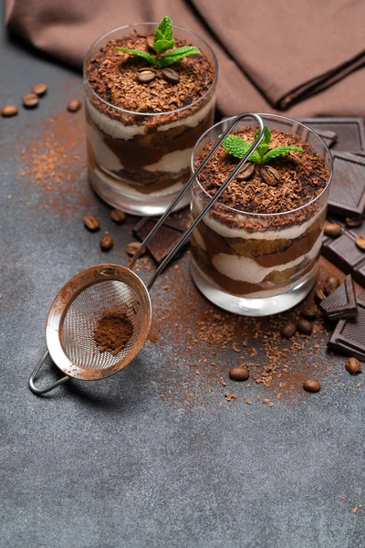 Klassisches Tiramisu-Dessert im Glasbecher, Sieb und Schokoladenstückchen auf dunklem Betongrund — Stockfoto