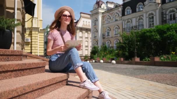 Schöne junge Frau Touristin Angenehm mit Stadtplan sitzt auf Treppen im Stadtzentrum — Stockvideo