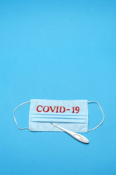 Mascarilla médica azul desechable con signo COVID-19 y termómetro electrónico sobre fondo azul —  Fotos de Stock
