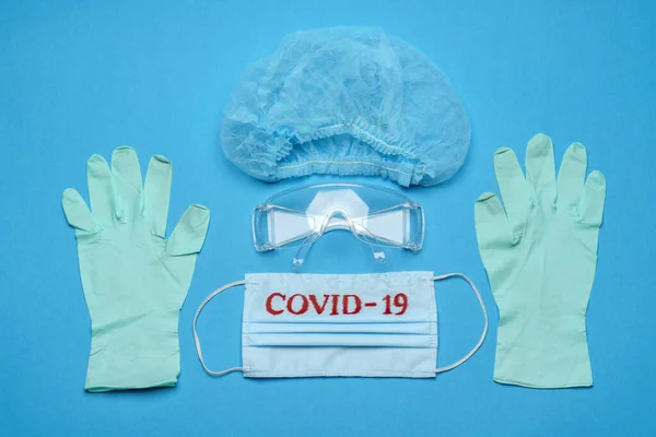 Mascarilla médica azul desechable con signo COVID-19, gafas protectoras, guante de látex de goma y sombrero sobre fondo azul —  Fotos de Stock