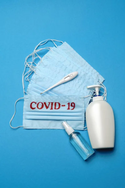 Pila de mascarillas médicas azules desechables con signo COVID-19 y desinfectante de manos con alcohol antiséptico sobre fondo azul —  Fotos de Stock