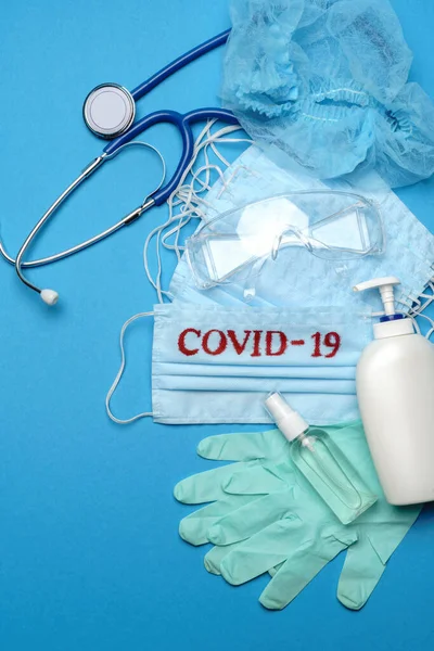 Pila di maschere monouso blu medico con segno COVID-19, guanti in lattice di gomma, occhiali, stetoscopio e alcol disinfettante mano antisettico su sfondo blu — Foto Stock