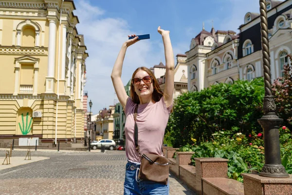 Mooie jonge vrouw toerist in het centrum van de stad met telefoon en het maken van ja win gebaar — Stockfoto