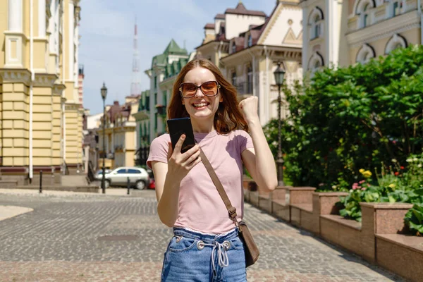 Belle jeune femme touriste dans le centre-ville tenant le téléphone et faisant oui geste de victoire — Photo