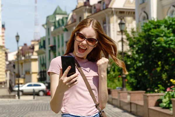 Hermosa joven turista en el centro de la ciudad sosteniendo el teléfono y haciendo sí ganar gesto —  Fotos de Stock