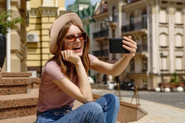 Piękna młoda kobieta turysta siedzi na schodach za pomocą smartfona podejmowania rozmowy wideo lub selfie — Zdjęcie stockowe