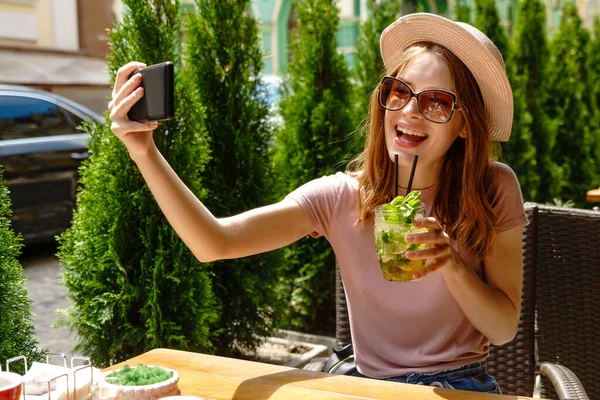 Młoda kobieta pije koktajl mojito na tarasie kawiarni w gorący letni dzień i robi zdjęcie selfie — Zdjęcie stockowe