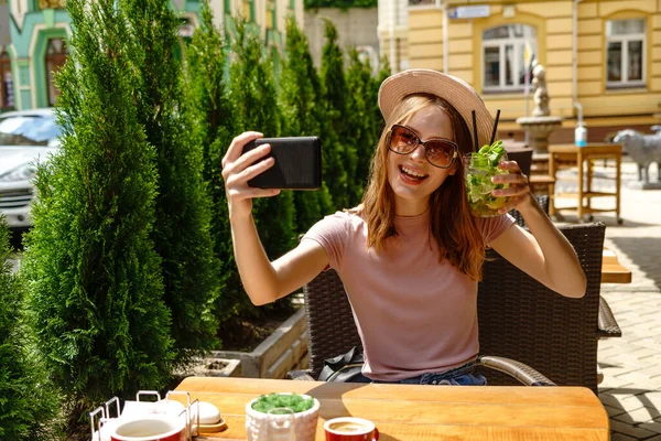 Mujer joven bebiendo cóctel mojito en la terraza de la cafetería en el caluroso día de verano y haciendo selfie shot —  Fotos de Stock