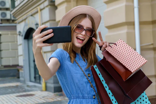 Młoda kobieta z torbami na zakupy robi selfie spacerując w mieście w letni dzień — Zdjęcie stockowe