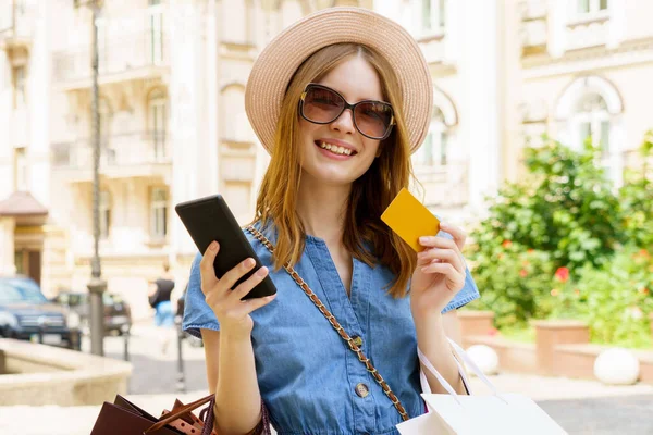 Молода жінка з сумками тримає телефон і картку, що йде в місті в літній день — стокове фото