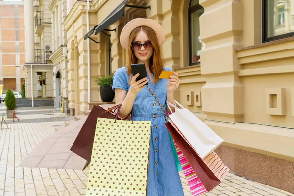 Mladá žena s nákupními taškami drží telefon a karty procházky ve městě v letní den — Stock fotografie