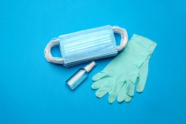 Pila di maschere mediche blu monouso, guanti in lattice di gomma e disinfettante mano alcol antisettico su sfondo blu — Foto Stock