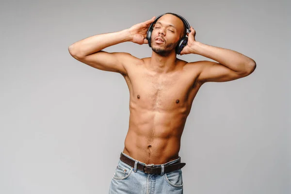 Africano americano jovem sem camisa ouvindo música em fones de ouvido sobre fundo cinza claro — Fotografia de Stock