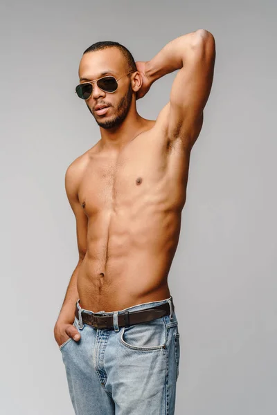 Сексуальний молодий м'язистий афроамериканський чоловік без сорочки носить сонцезахисні окуляри на світло-сірому фоні — стокове фото