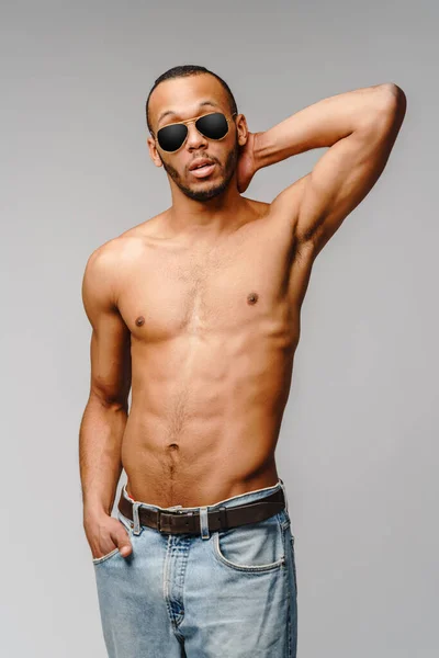 Сексуальний молодий м'язистий афроамериканський чоловік без сорочки носить сонцезахисні окуляри на світло-сірому фоні — стокове фото