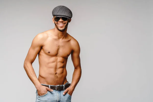 Σέξι νεαρός μυώδης Αφροαμερικάνος χωρίς πουκάμισο φορώντας γυαλιά ηλίου και καπέλο πάνω από ανοιχτό γκρι φόντο — Φωτογραφία Αρχείου