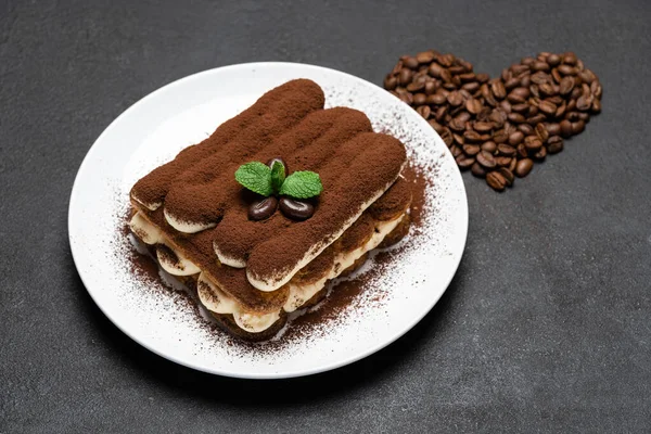 Porção de sobremesa tiramisu clássico na placa de cerâmica e grãos de café em forma de coração no fundo de concreto — Fotografia de Stock