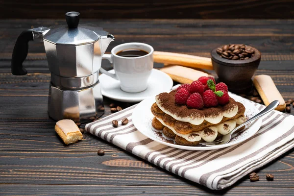 Часть классического десерта тирамису с малиной, чашкой эспрессо и кофеваркой на деревянном фоне — стоковое фото
