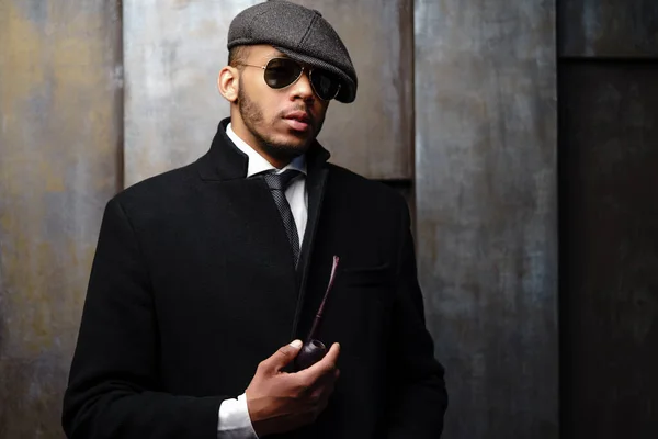 Studio shot of stylish african american man wearing coat, cap, glasses and holding smoking pipe — Stock Photo, Image