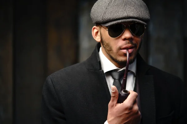 Studio shot di elegante uomo afroamericano indossando cappotto, berretto, occhiali e tenendo la pipa fumante — Foto Stock