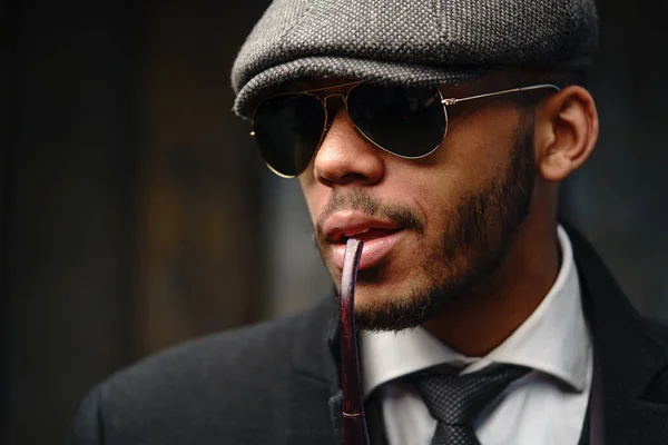 Studio shot of stylish african american man wearing coat, cap, glasses and holding smoking pipe — Stock Photo, Image