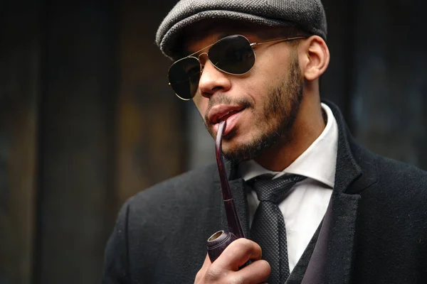 Studio shot of stylish african american man wearing coat, cap, glasses and holding smoking pipe — Stock Photo, Image
