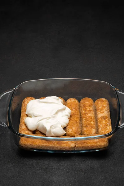 Tiramisu sobremesa cozinhar - Tradicional italiano Savoiardi ladyfingers Biscoitos e creme em assadeira de vidro no fundo de concreto escuro — Fotografia de Stock