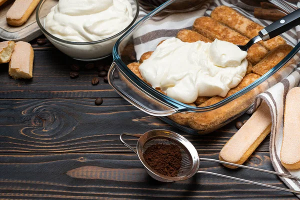 Tiramisu dessert cooking - Traditional Italian Savoiardi ladyfingers Biscuits and cream in glass baking dish and coffee on wooden background — Stock Photo, Image