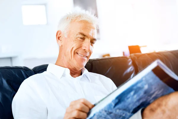Bonito senior homem lendo um livro relaxante em um sofá — Fotografia de Stock
