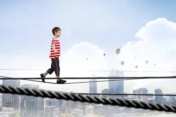 Cute joyful little boy walking on a rope over city. Mixed media — Stock Photo, Image