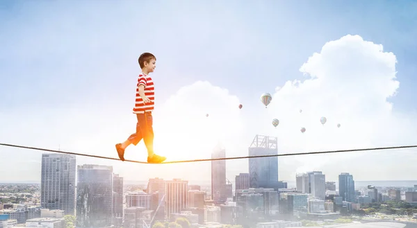 Schattige vrolijke kleine jongen lopen op een touw over stad. Mixed media — Stockfoto
