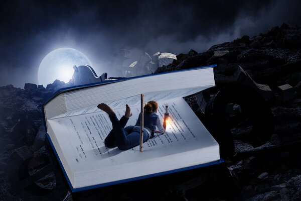 Little woman lying with a flashlight in a large book opened with wooden natural bookmark