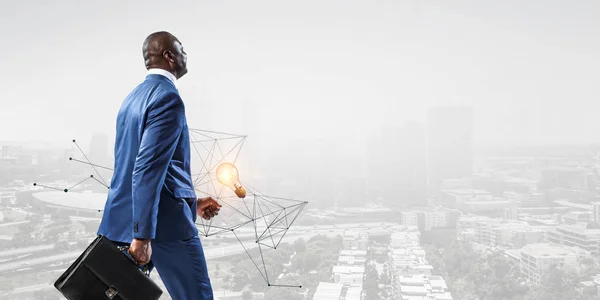 Back view of smiling black businessman with a briefcase on a cityscape background with 3d graph and a burning lamp — Stock Photo, Image