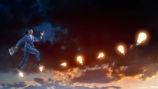 Hombre de negocios negro corriendo sobre hipotéticas escaleras hechas de lámparas encendidas sobre un oscuro cielo nublado de noche —  Fotos de Stock