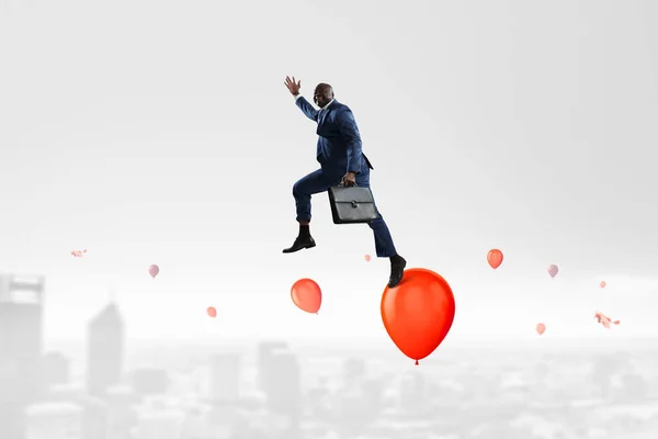 Zwarte lachende zakenman springt over rode ballonnen in de lucht boven stedelijk gebied — Stockfoto