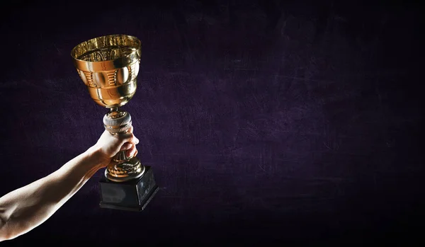 Hand holding up a gold trophy cup against dark background — Stock Photo, Image