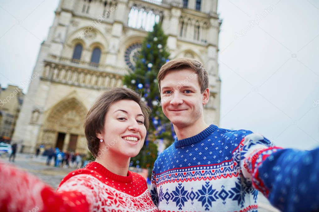 Happy couple in colorful sweaters taking selfie with mobile phone near Notre-Dame cathedral and decorated Christmas tree in Paris, France