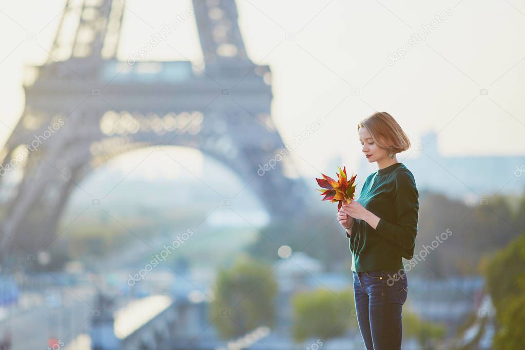 Beautiful young French woman with bunch of colorful autumn leaves near the Eiffel tower in Paris on a fall day