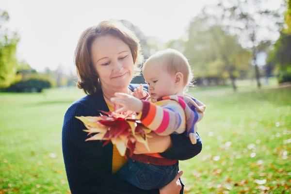 愉快的微笑的妇女与女婴和一堆红色枫叶在阳光明媚的秋天天在公园 秋季活动与孩子 — 图库照片