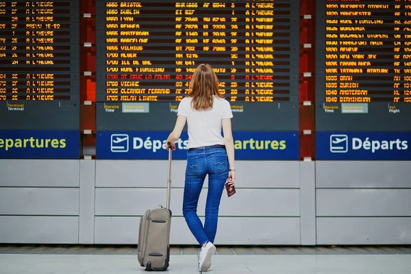Young woman in international airport with luggage and passport near flight information display