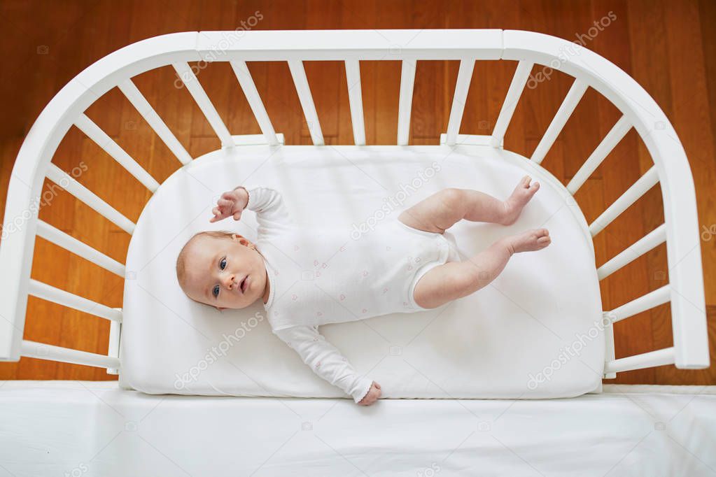 Adorable baby girl in co-sleeper crib attached to parents' bed. Little child having a day nap in cot. Infant kid in sunny nursery