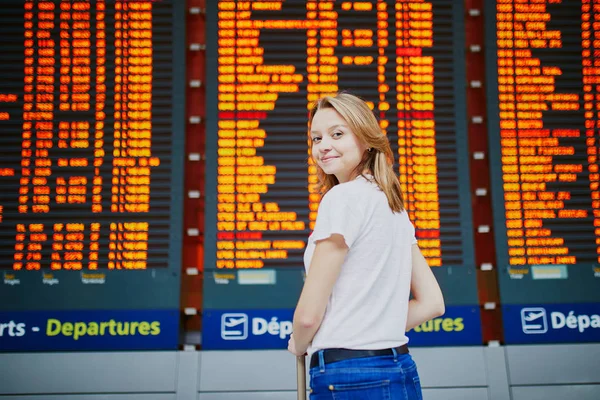 Young woman in international airport with luggage near flight information display