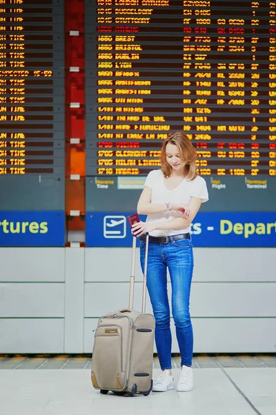Young woman in international airport with luggage near flight information display, looking at her watch to check time. Delayed or cancelled flight concept