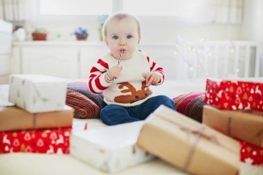 Sıcak bir Noel süveteri giyip Noel hediyelerini ilk Noel 'inde açan mutlu küçük bir kız bebek. Evde çocuklarla Noel 'i kutluyoruz.