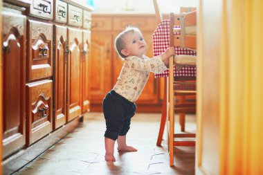 Baby girl standing on the floor in the kitchen and holding on to furniture. Little child oulling up at home clipart